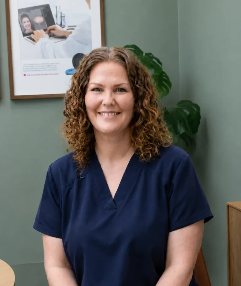 Smiling female healthcare professional with curly hair in navy scrubs, seated indoors with framed posters behind her.