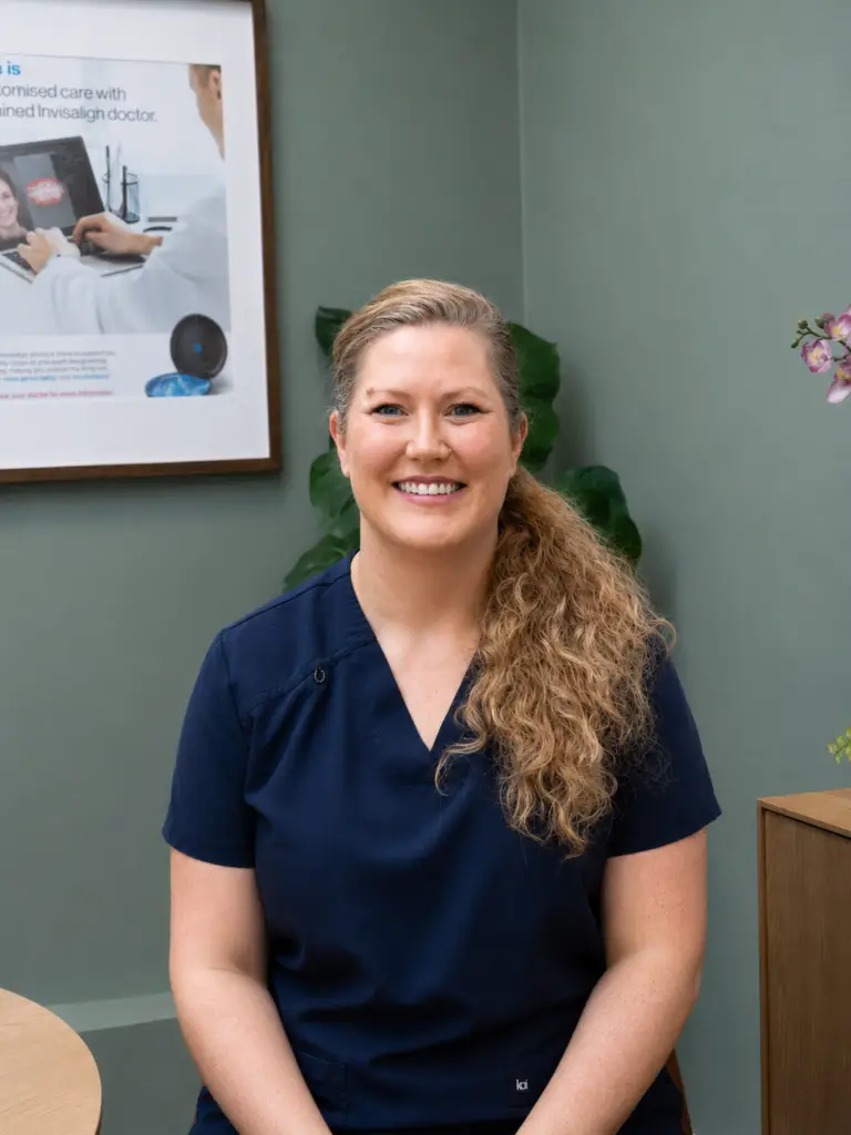 Portrait of a smiling woman in navy scrubs seated in a clinic setting with a poster on the wall behind her.